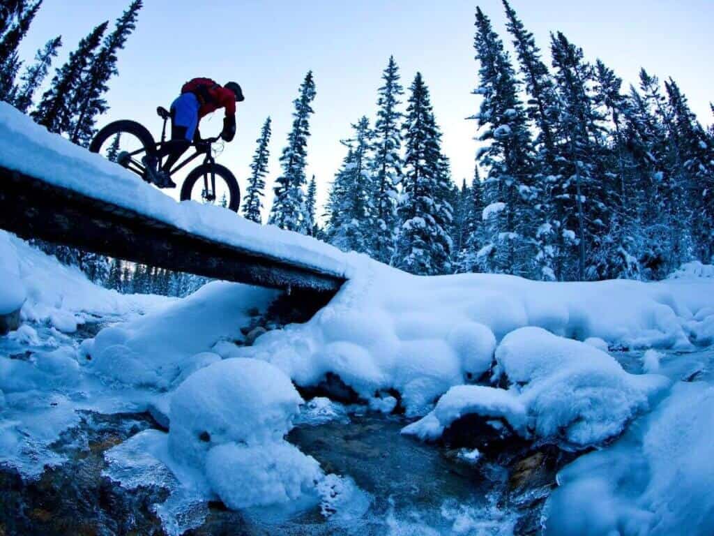 Person riding fat bike over snowy foot bridge with creek running underneath and snowy winter landscape around