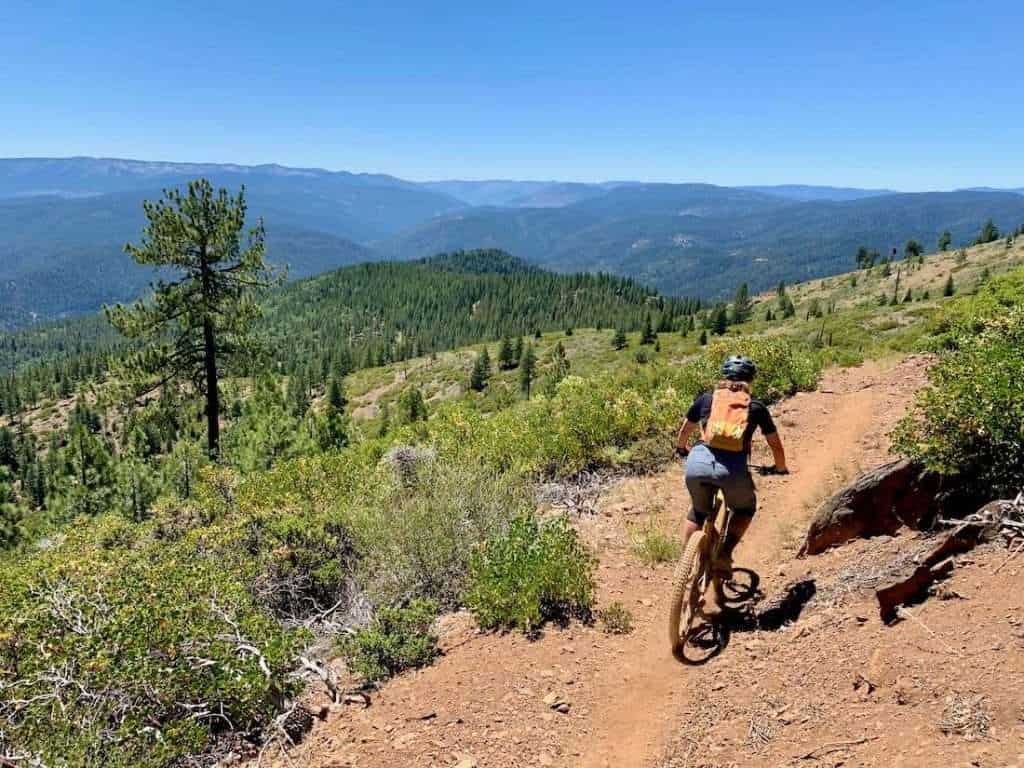 Female mountain biker riding on scenic section of singletrack trail with expansive views of rolling forested mountain range