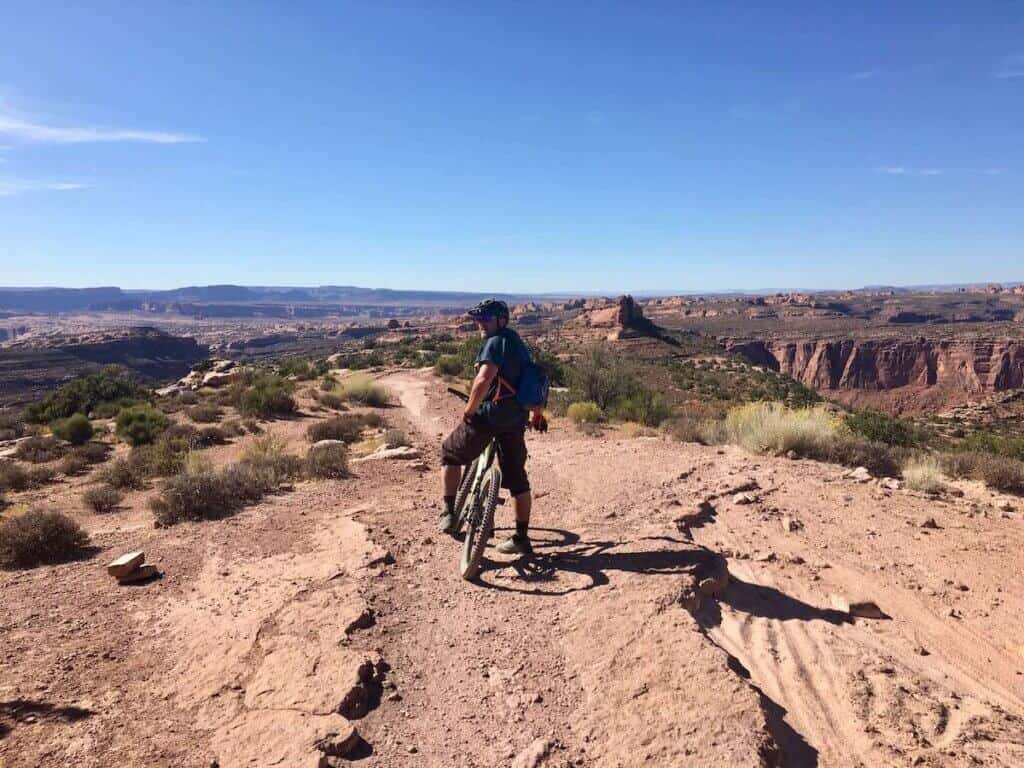 Man on mountain bike looking back over shoulder at camera on The Whole Enchilada Trail in Moab Utah with canyonland views in background