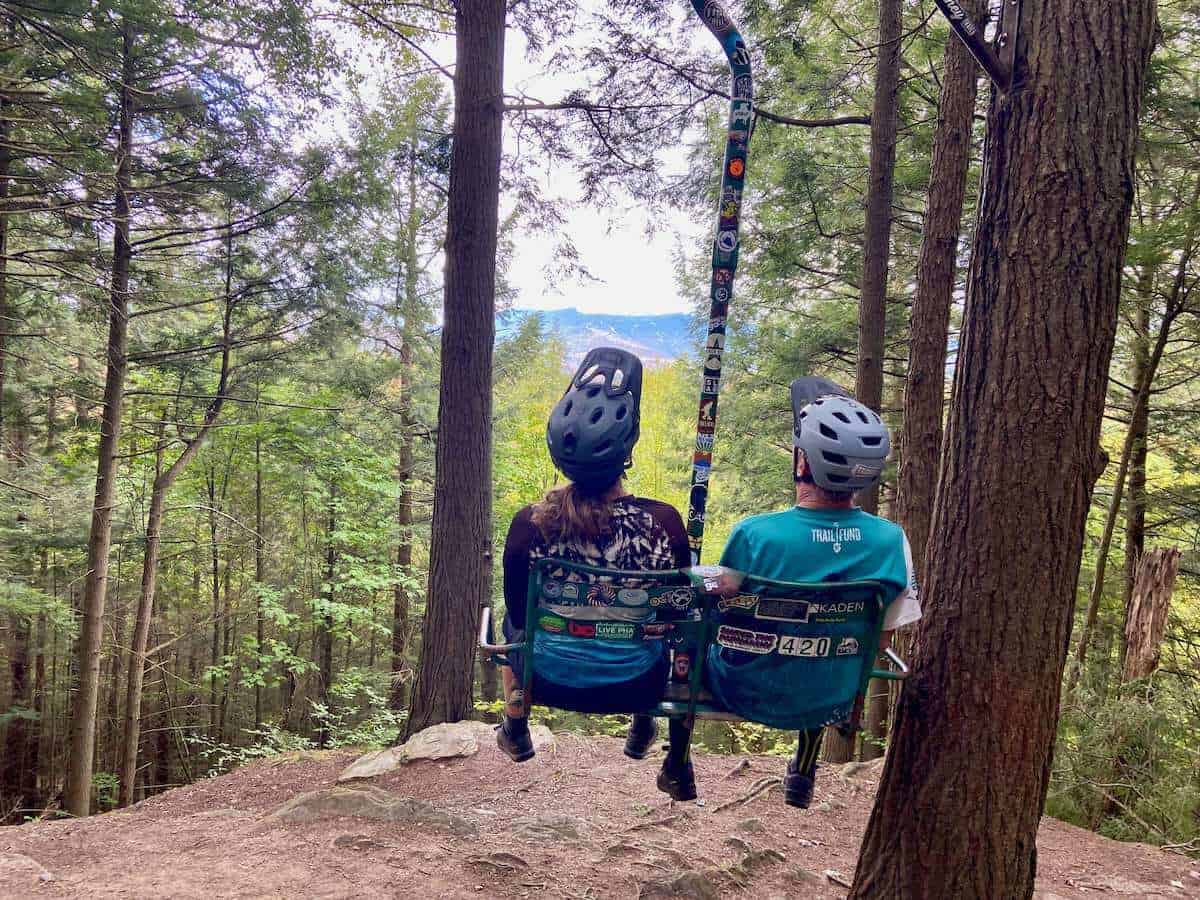Two people sitting on chairlift on mountain bike trail facing away from camera with helmets on