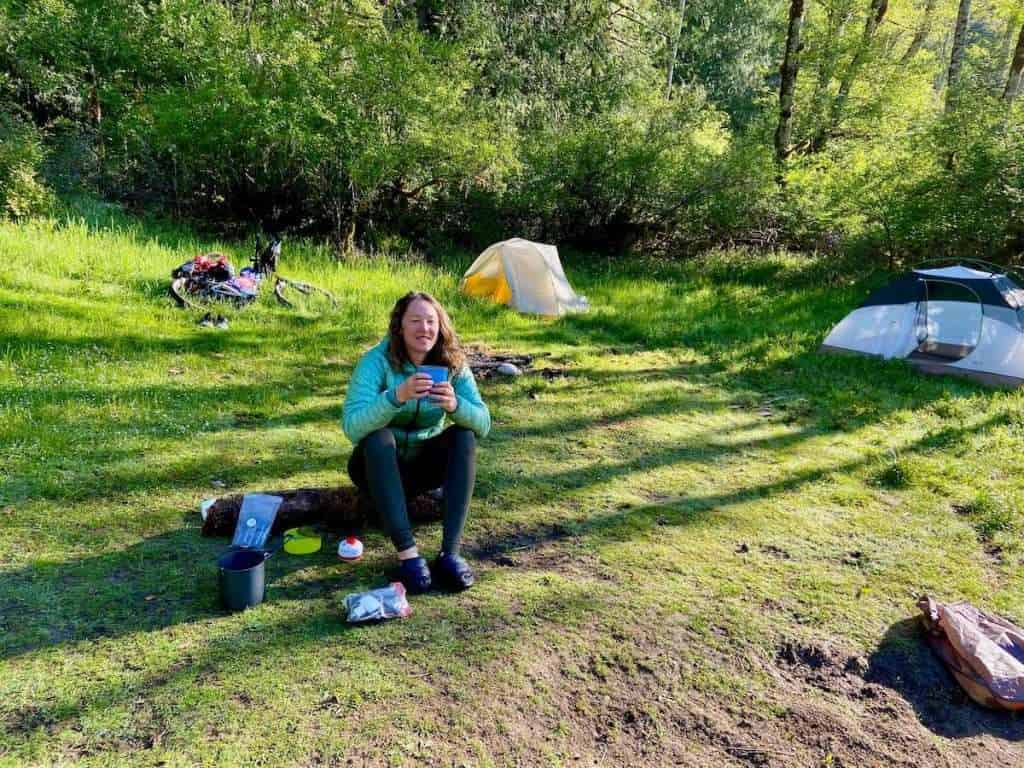Woman sitting on grass at dispersed campsite sipping coffee from mug with camp gear strewn about