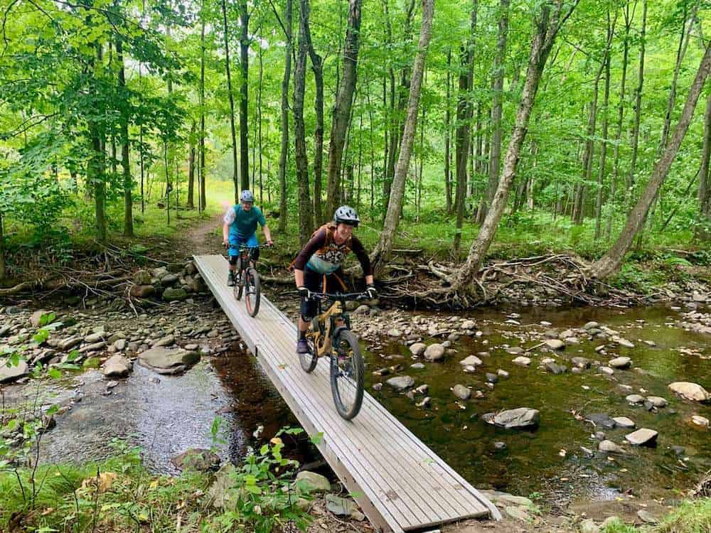 Two mountain bikers riding across narrow bridge over shallow river in lush green forest of Vermont