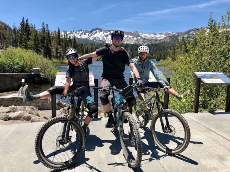 Three mountain bikers posing for camera holding each others shoulders while balancing on bikes with mountains and lakes in background