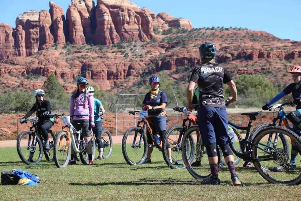 Group of mountain bikers standing next to bikes listening to coach talk on grassy field with Sedona's red rock bluffs in background