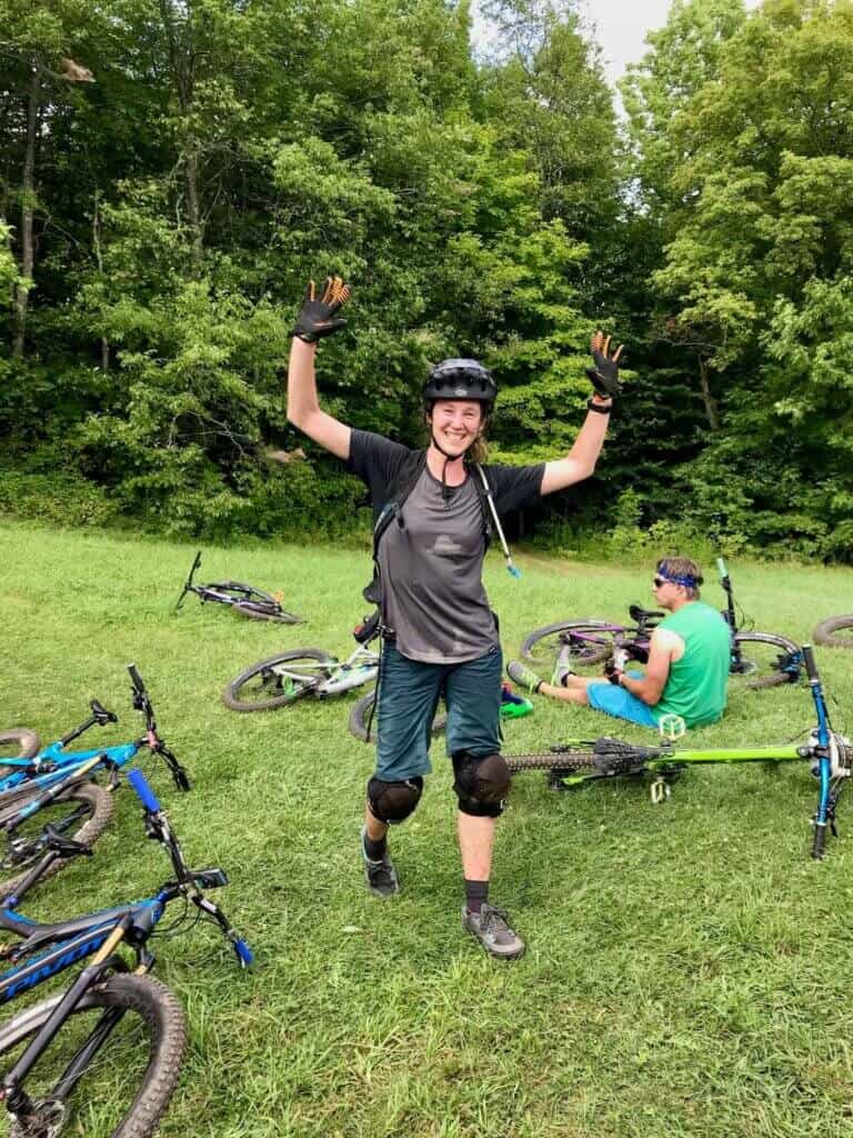 Becky walking toward camera with hands up in the air and smiling. Clothes are drenched in sweat and mountain bikes laying on ground around her