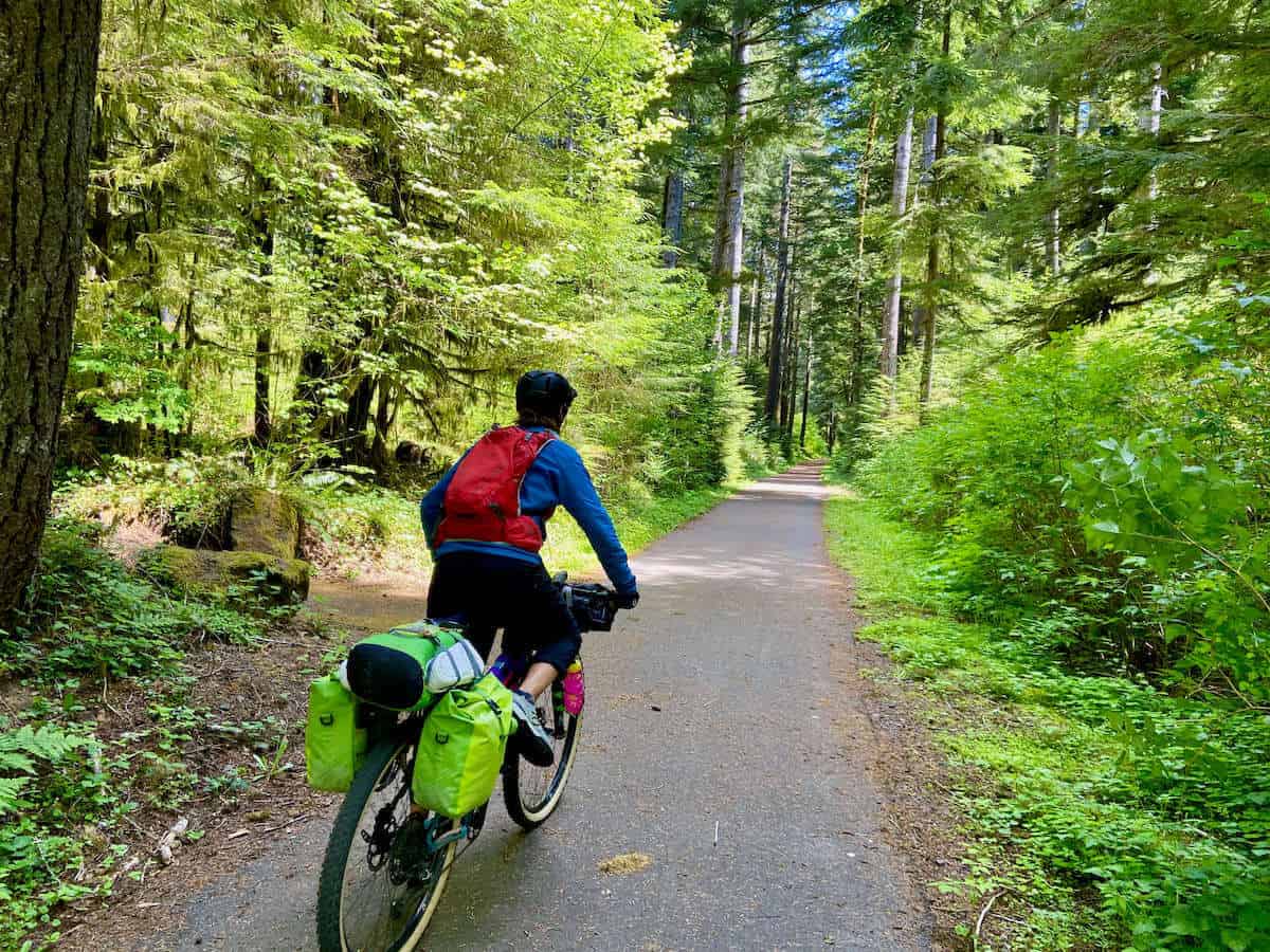 Cyclist with bike loaded with gear pedaling down paved bike path lined with lush trees and vegetation on the Olympic Peninsula in Washington