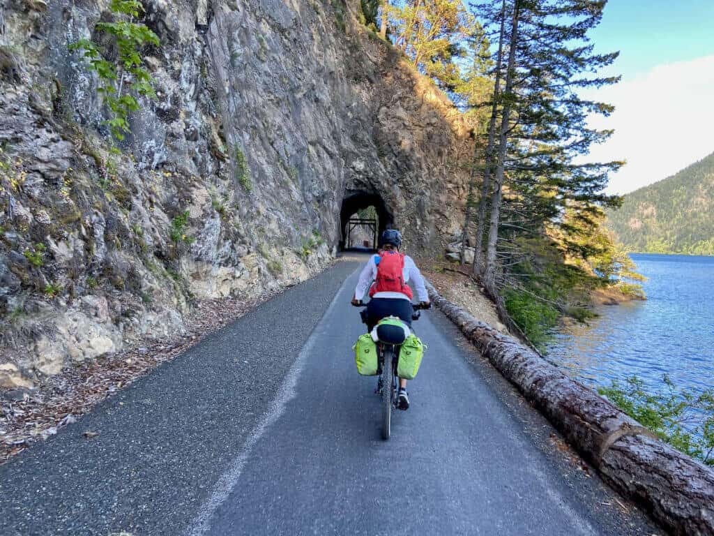 Cyclist riding on paved bike path next to lake about to ride under tunnel through rock