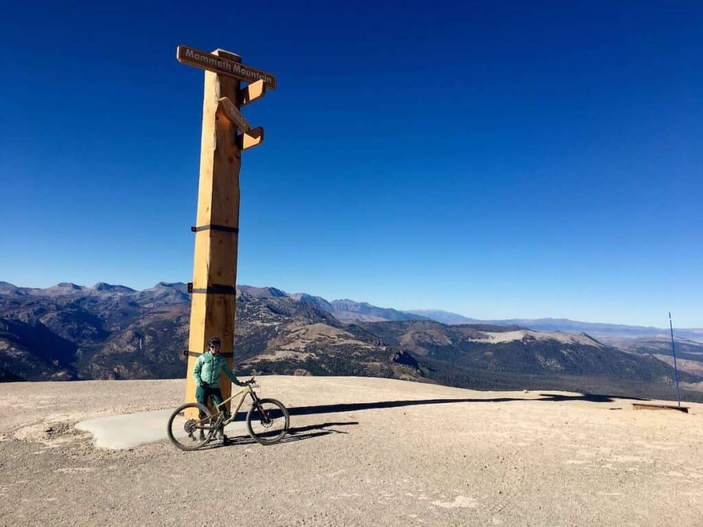 Woman standing next to bike and tall wooden sign at the top of Mammoth Mountain in California
