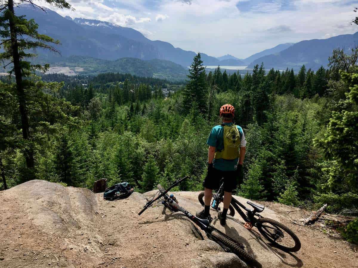 Mountain biker standing next to bikes on rock slab lookout overlooking mountains and fjord in Squamish, British Columbia
