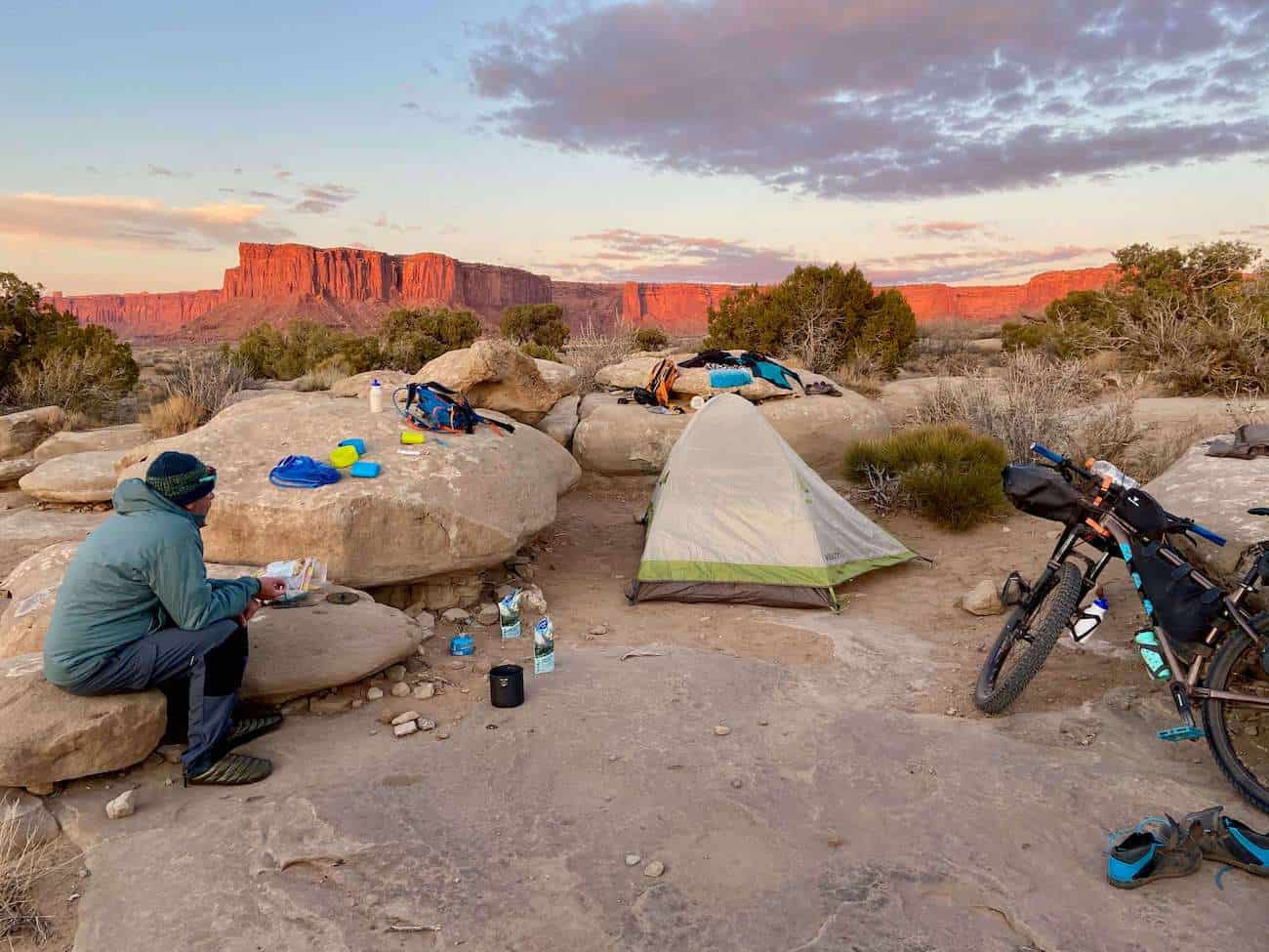 Man sitting on rocks at dispersed campsite with camping gear laid out around him and tent set up. Bikepacking bike learning against a large rock and sun setting on red rock bluffs in background
