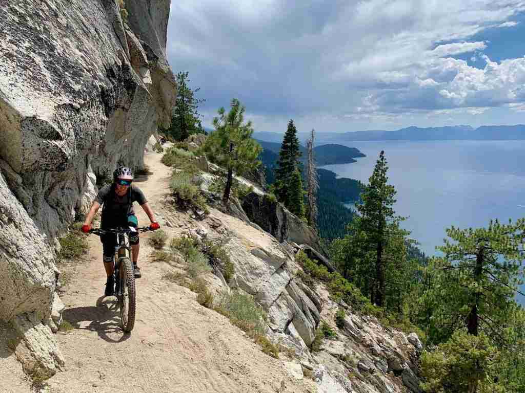 Becky riding a mountain bike along narrow cliffside singletrack trail in California with expansive views of Lake Tahoe behind her. 