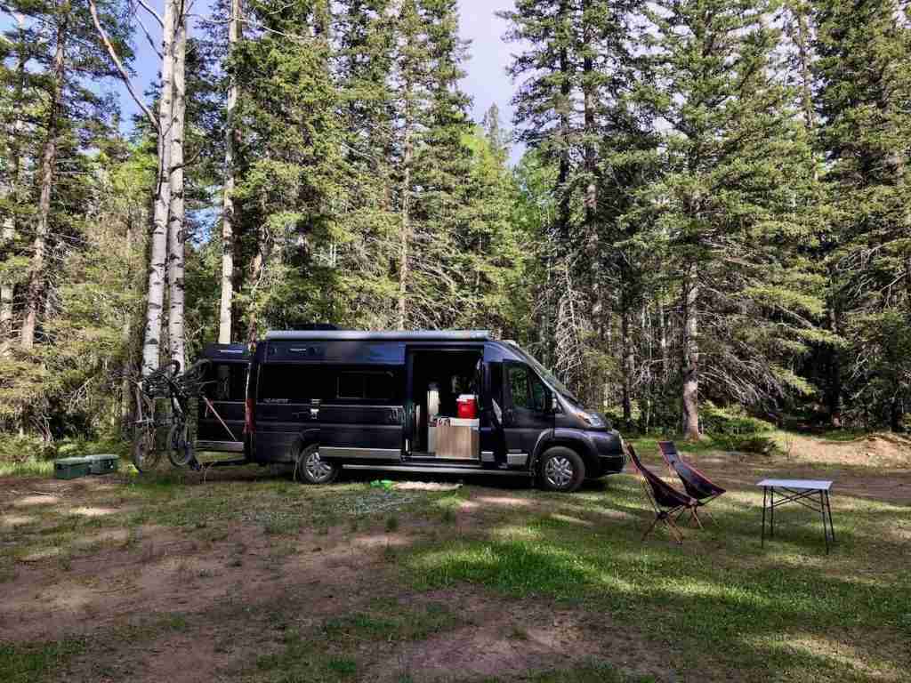 Van parked in grassy clearing with woods behind it. Table and chairs set out in frong