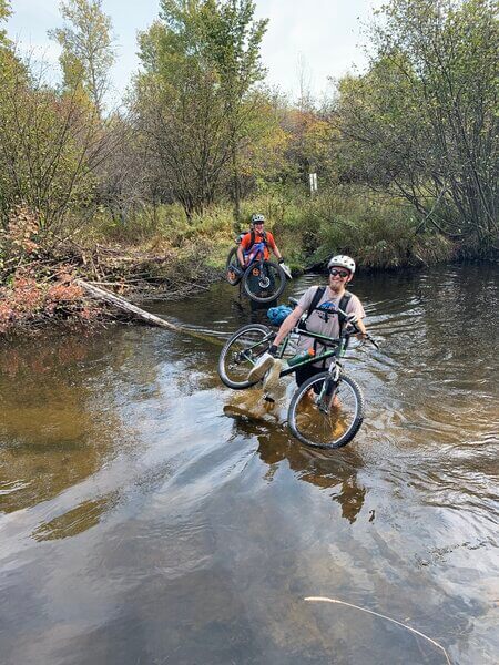 Two mountain bikers carrying bikes through river in Michigan