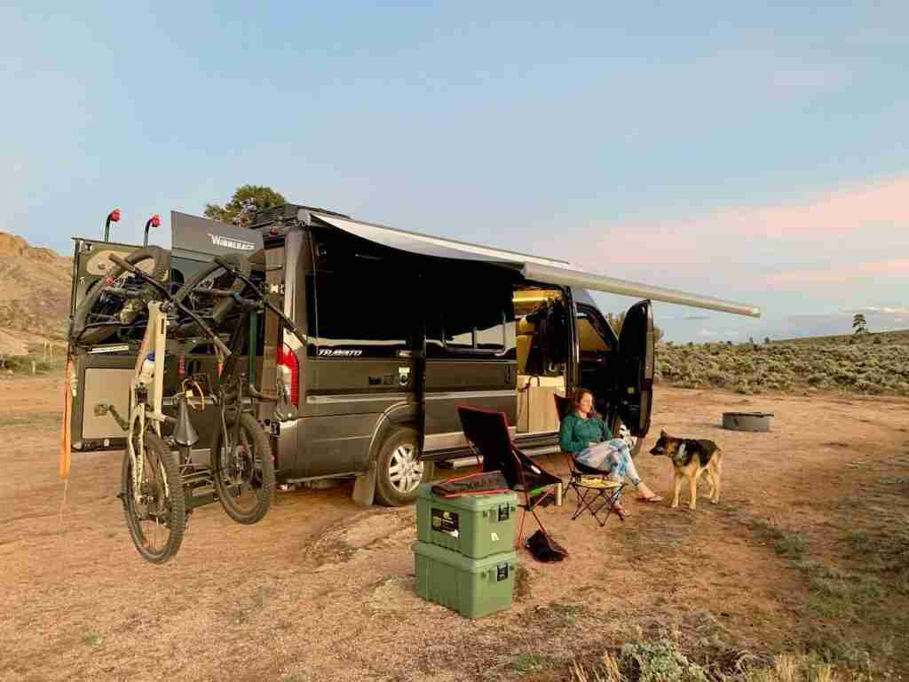 Woman sitting on chair outside of camper van with dog at her side. Bikes hanging from back of van