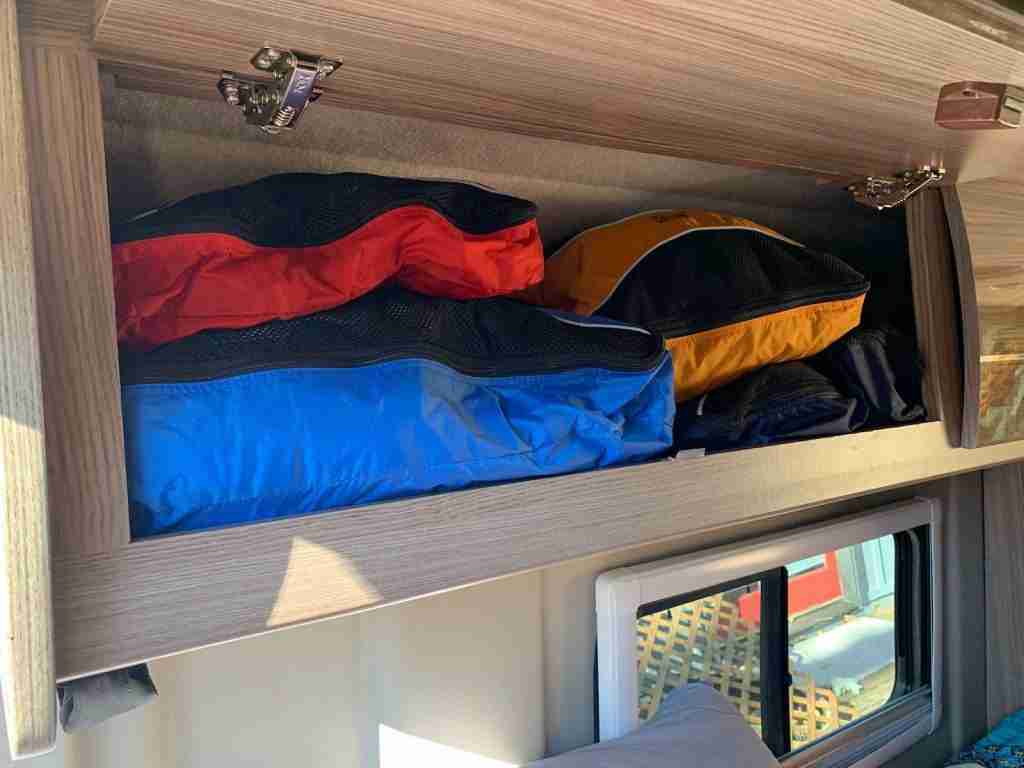 Packing cubes filled with clothes in cabinet over bed in converted camper van
