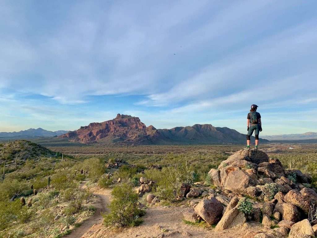 Woman mountain biker dressed in gear standing on rock pile next to trail looking out over stunning Phoenix, Arizona desert landscape
