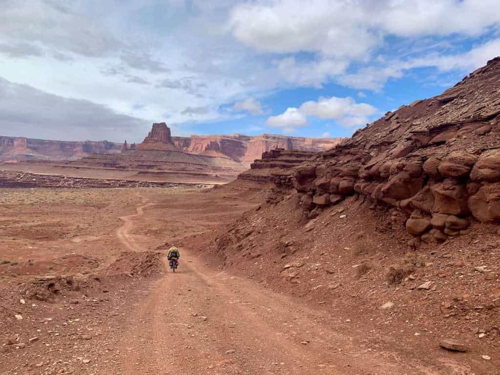 Bikepacker riding bike down remote red dirt road on the White Rim Trail in Utah with canyonland vistas in the distance