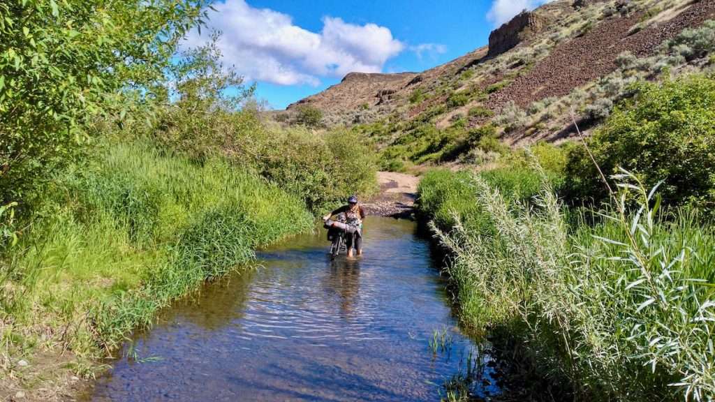 Woman pushing loaded bikepacking bike through flooded section of remote dirt road in eastern Washington