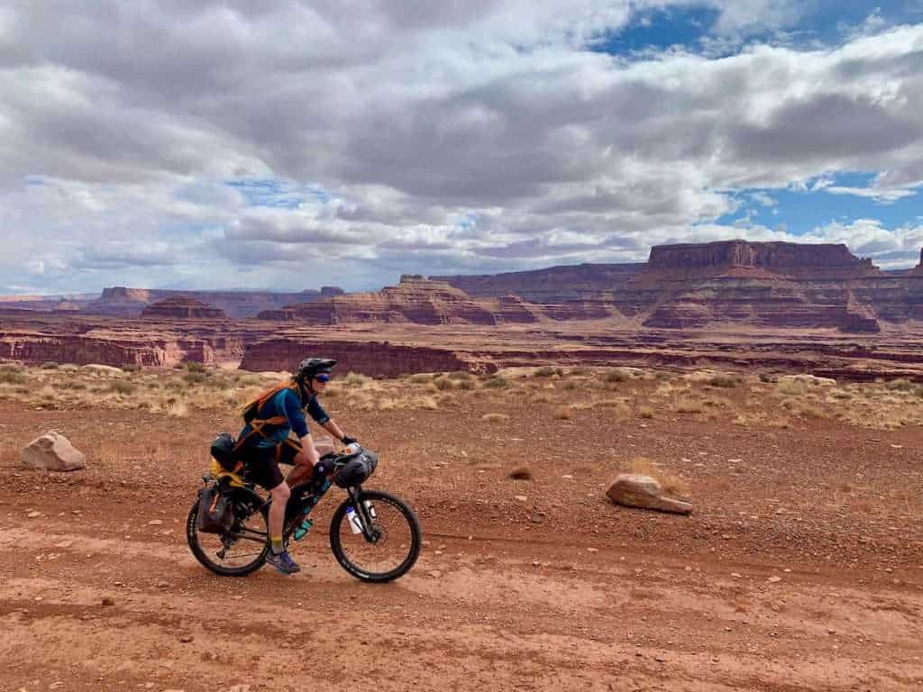 Becky on loaded bikepacking bike on White Rim Trail in Canyonlands National Park with tall red bluffs in the background