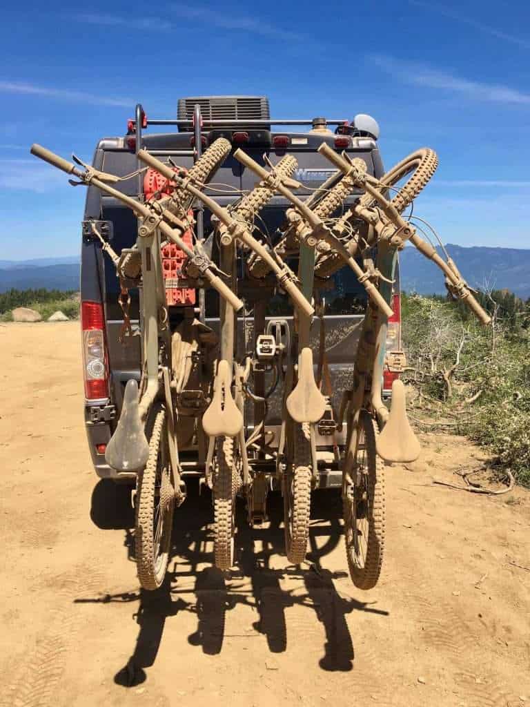 Four mountain bikes on a hanging rack on the back of a van completely covered in dust