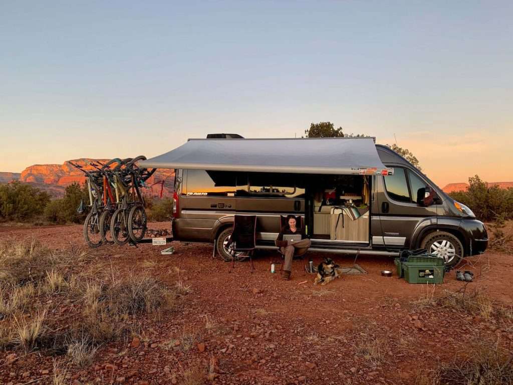 Becky sitting in camp chair in front of van with sliding door open and awning out. Four mountain bikes hang on bike rack behind van.