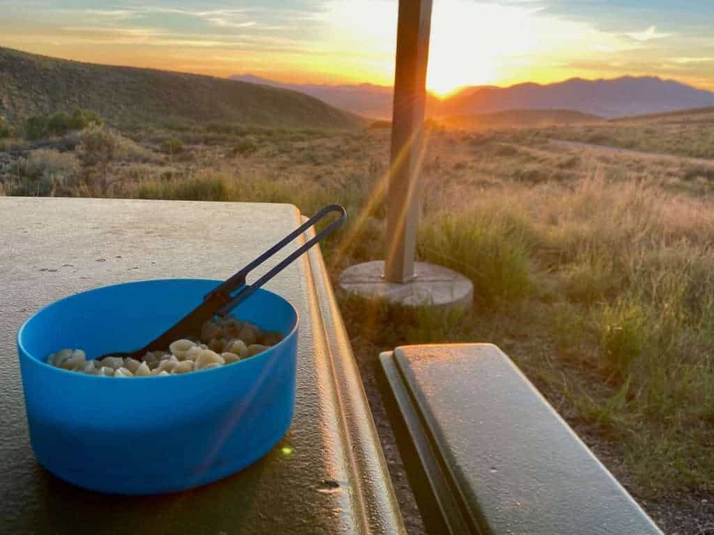 Bowl of pasta on picnic table with setting sun going down behind mountains in the background