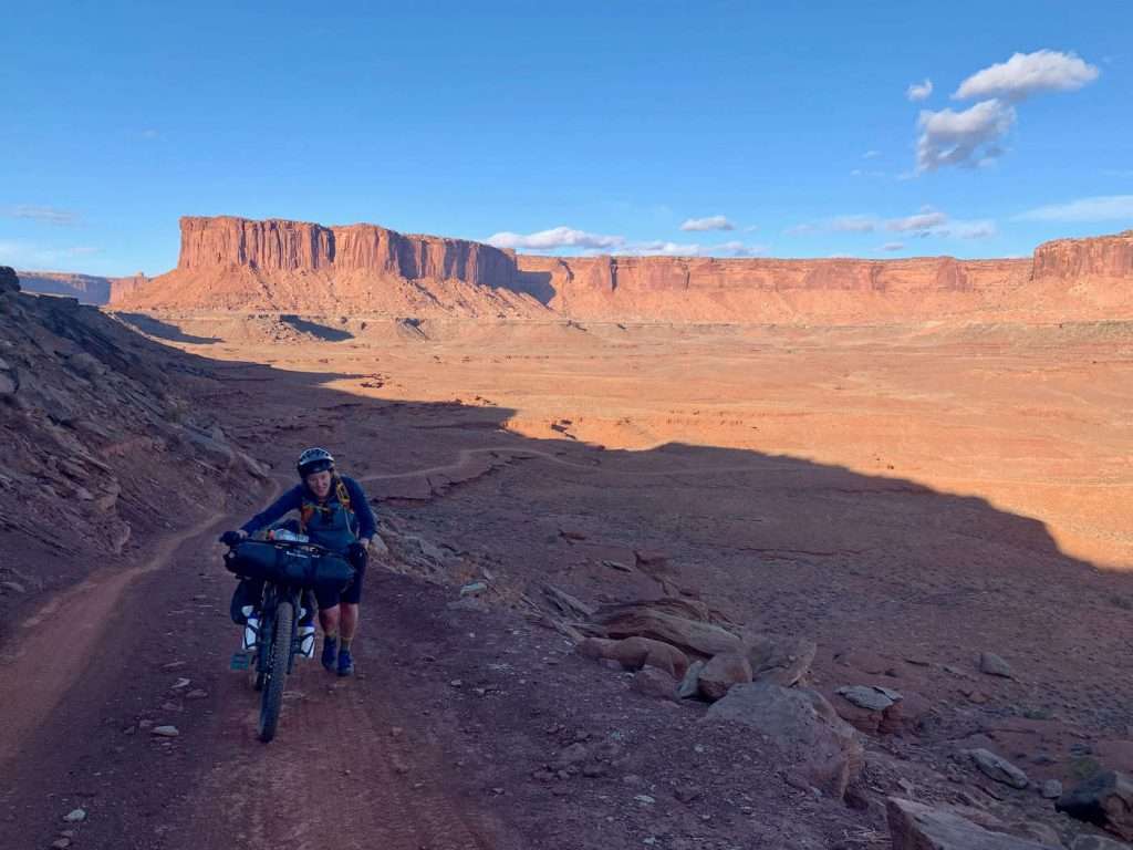 Becky pushing heavy loaded bikepacking bike up steep gravel road on White Rim Trail in Utah. She is in shadow while red rock bluffs in background are illuminated