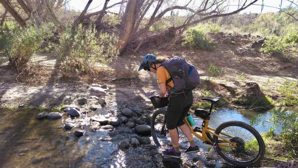 Female bikepacker walking bike across narrow and shallow river