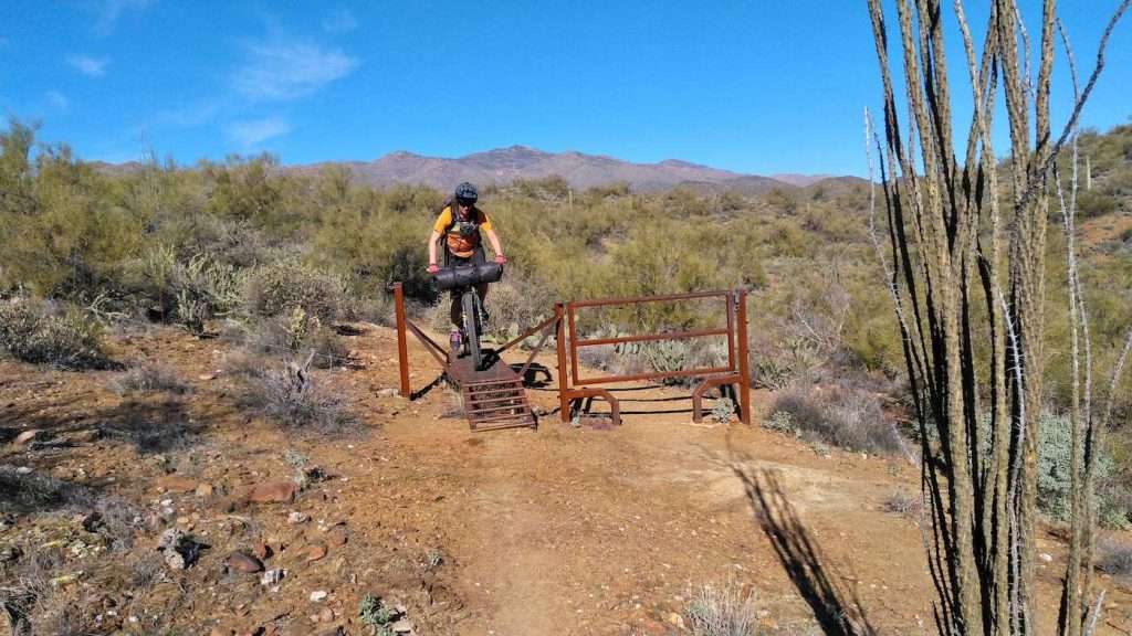 Female mountain biker with bikepacking set up in the desert riding over narrow metal ladder bridge, which acts as a cattle guard.