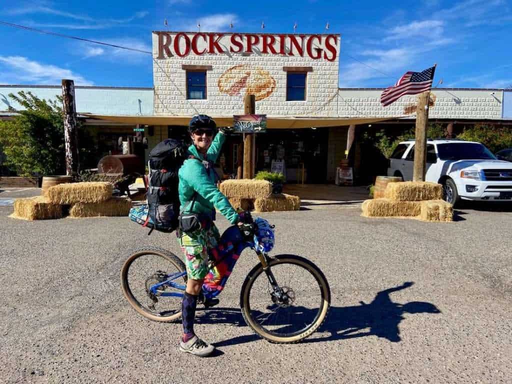 Female bikepacker straddling fully loaded bike in front of Rock Springs cafe. Rider is pointing at sign