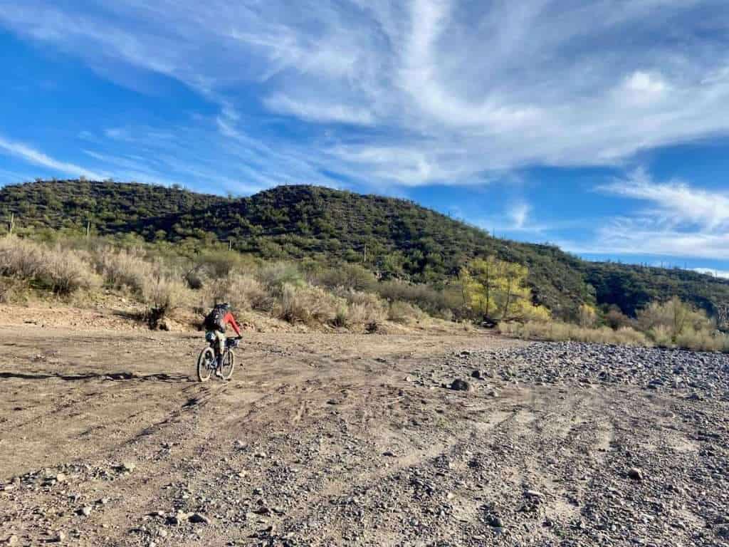 Female mountain biker crossing dry, sandy river bed in Arizona with vegetation-covered hill in the background.