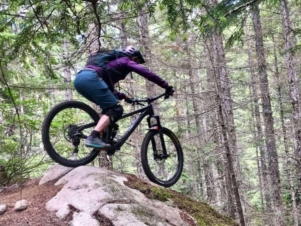 Female mountain biker riding over large rock slab on bike in pine forest. 
