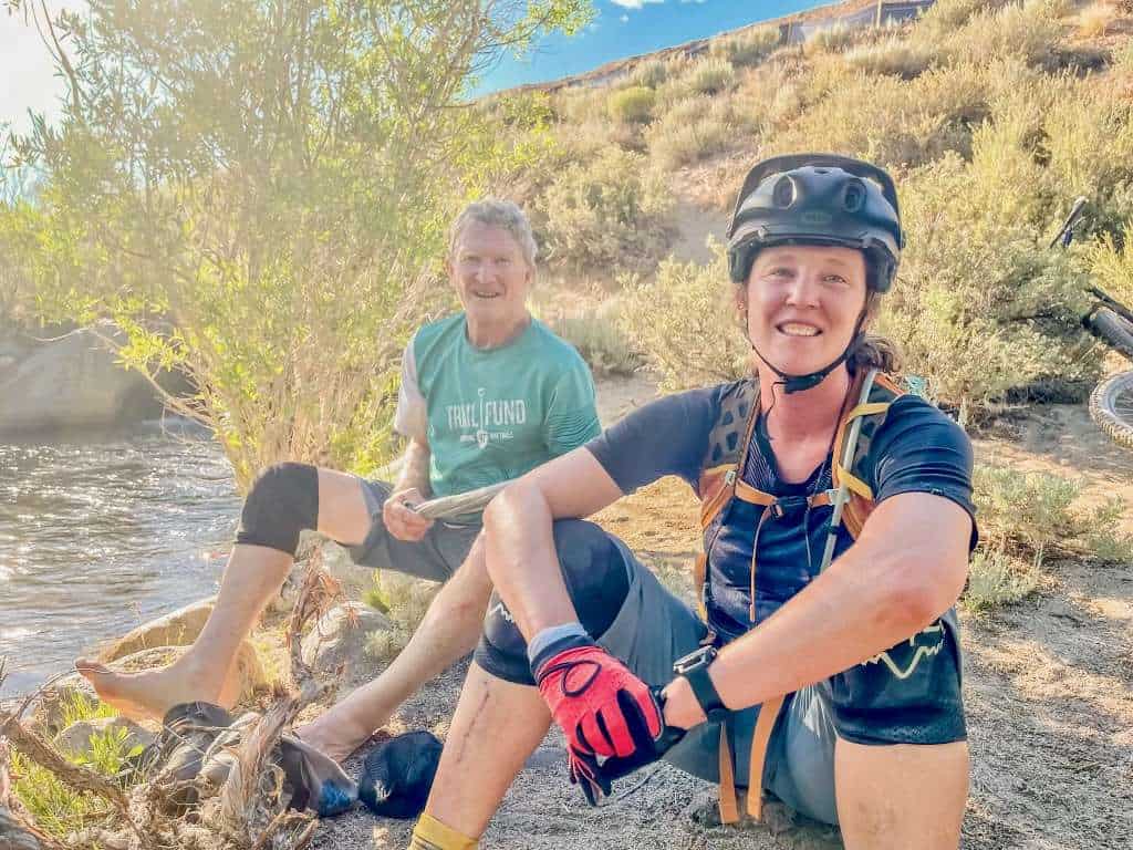 Becky Timbers, founder of Two Wheeled Wanderer, sitting on ground next to river wearing mountain bike gear, smiling at camera. Dad is behind her
