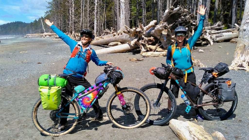 Two female mountain bikers posing behind loaded bikepacking bikes on beach in Washington at start of cross-washington mountain bike trip