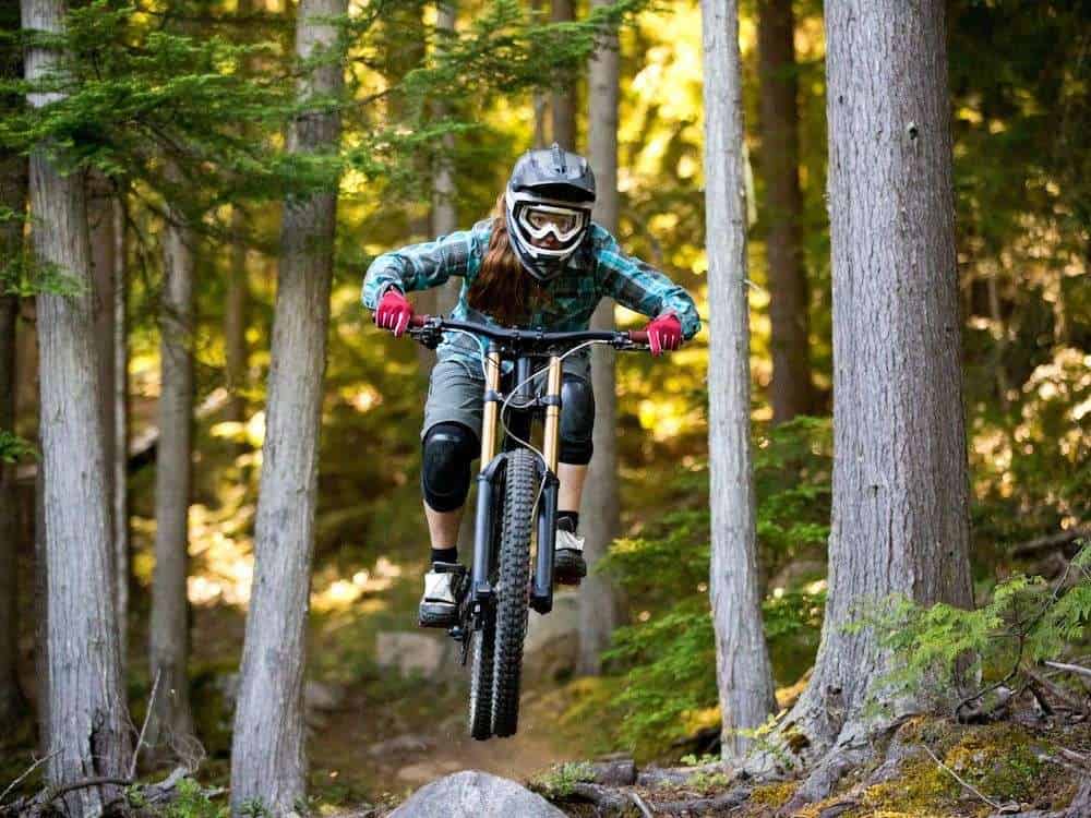 Female mountain biker wearing full-face helmet and goggles riding downhill bike through woods and jumping over rock 