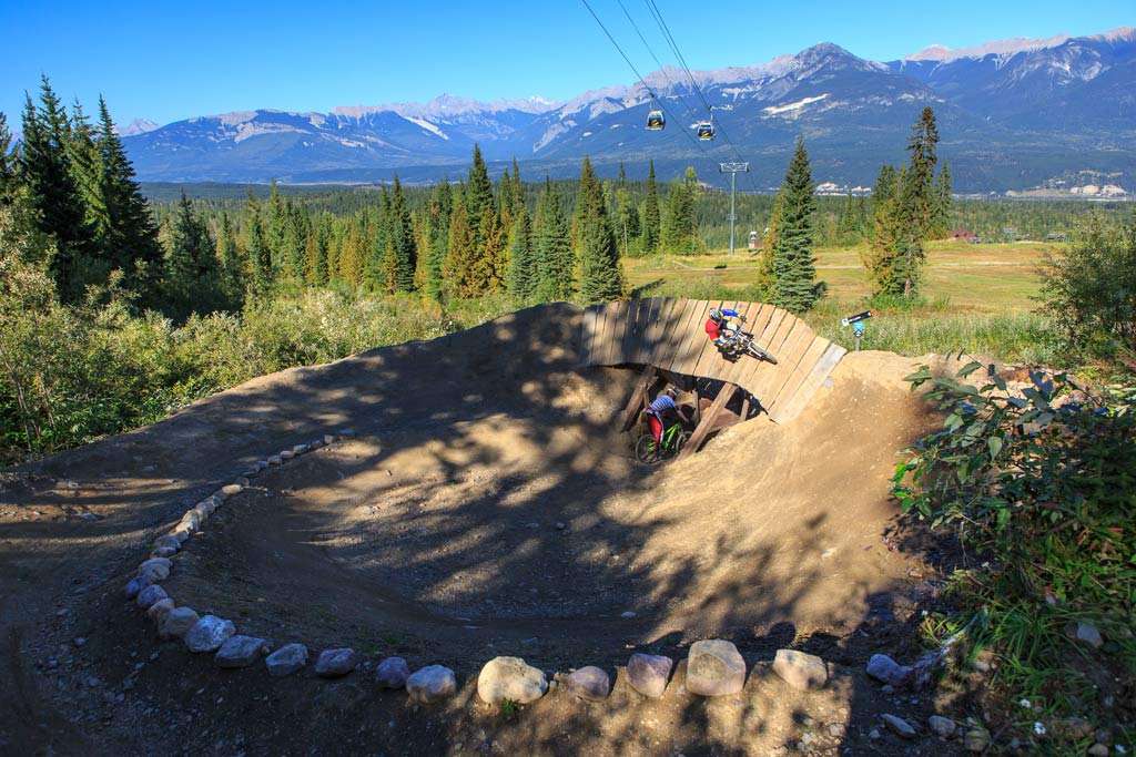 Two mountain bikers on downhill trail, one going over wooden wall ride, at Kicking Horse Bike Park in British Columbia. 