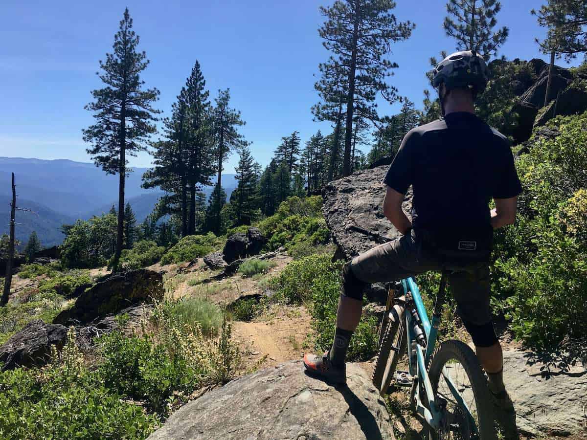 Mountain biker stopped on trail with foot placed on rock for balance facing away from camera.