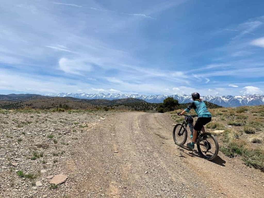 Becky riding bike to the summit of dirt road with tall snow-capped peaks in the distance