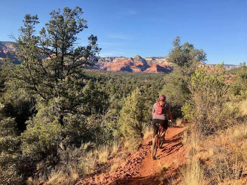 Mountain biker pedaling along singletrack trail in Sedona, Arizona with red rock bluffs in the distance
