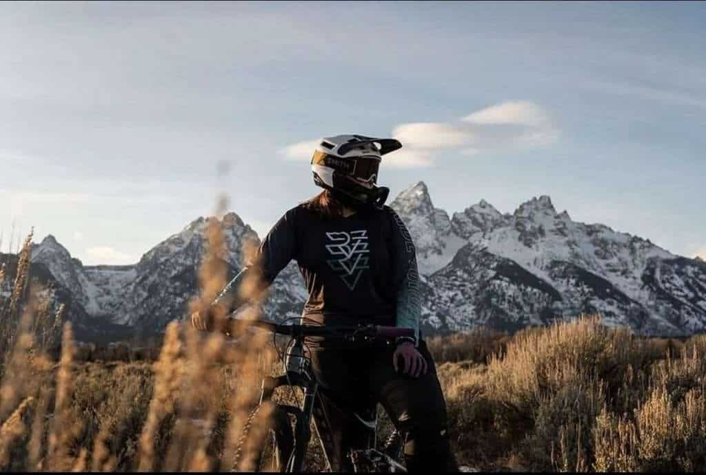 Female mountain biker in full mountain bike gear including full face helmet, goggles, and bike jersey. Snow-capped mountains in background