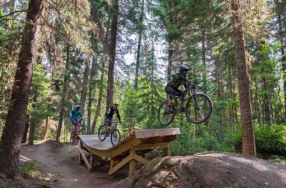 Three mountain bikers riding over wooden ramp and drop at Valemount bike park in British Columbia