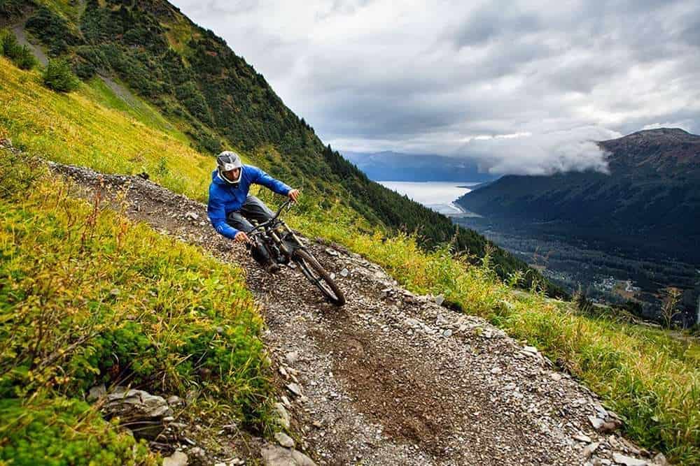 Mountain biker riding down bike park trail in Alaska with fjord and mountains in background