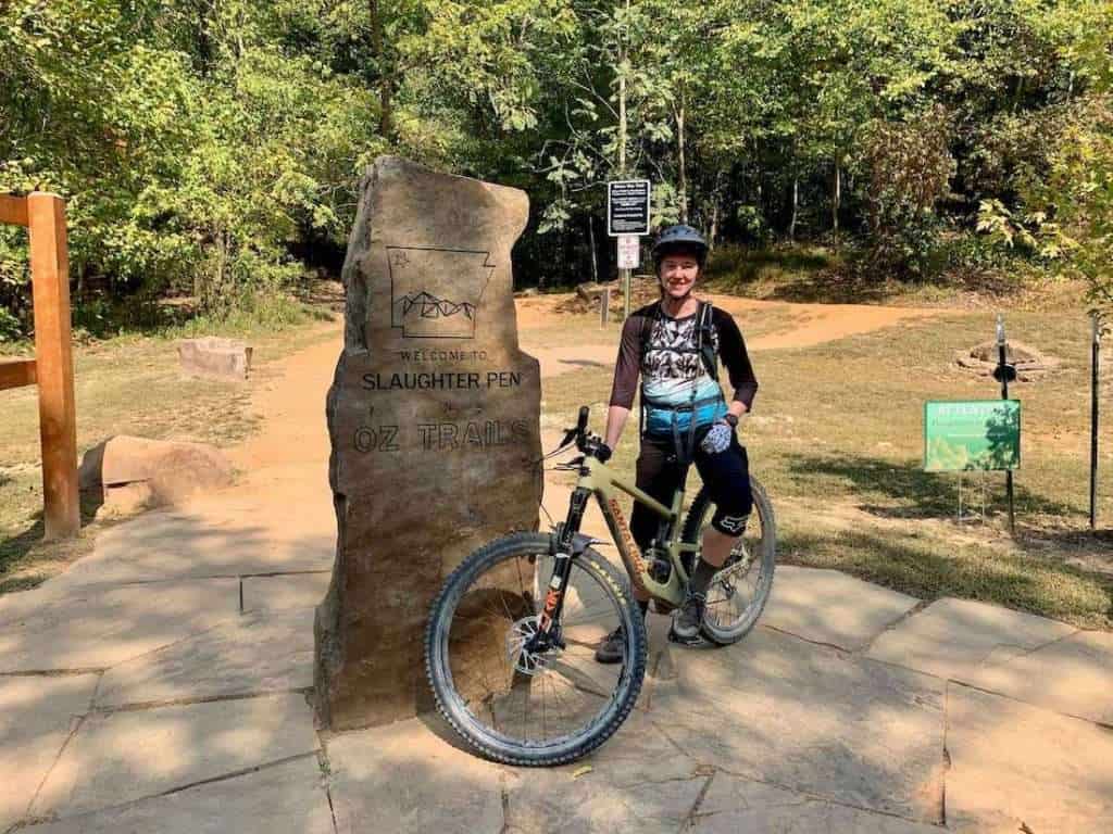 Becky standing over mountain bike next to rock slab sign that says "welcome to Slaughter Pen - Oz Trails"