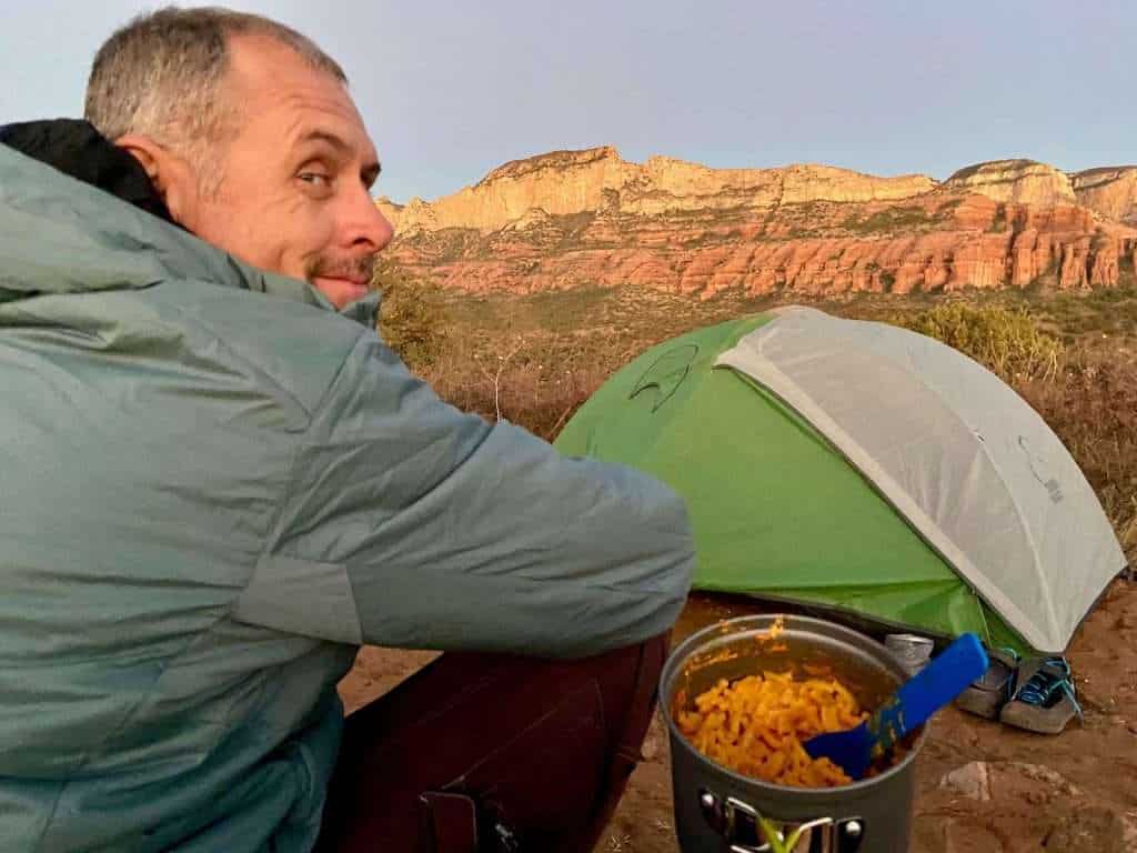 Man looking back at camera at campsite with pot of macaroni and cheese at his side. Views out over red rock bluffs of Sedona