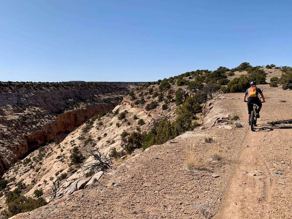 Becky riding mountain bike away from camera on cliff-side trail in Utah