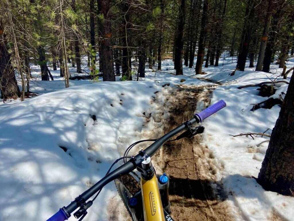 Photo out over front of mountain bike handlebars onto snowy and muddy mountain bike trail