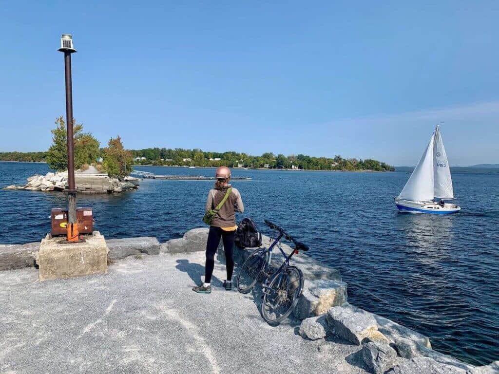 Becky standing next to bike on rock pier at The Cut in the Colchester Causeway on Lake Champlain with sailboat passing by