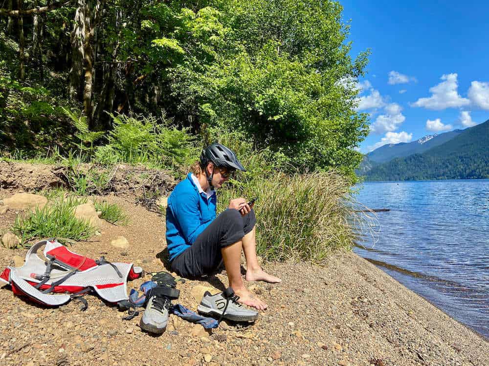 Cyclist sitting on sandy beach next to lake with shoes off and gear next to her