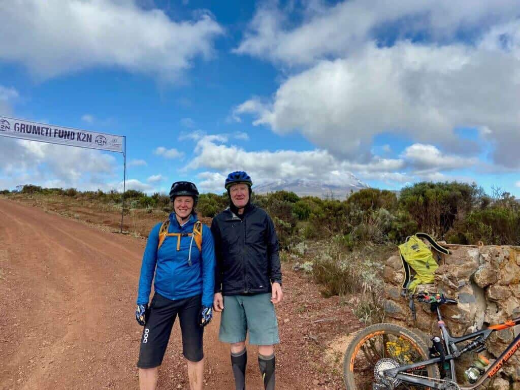 Becky and Mike in mountain bike gear standing in front of Grumeti Fund K2N banner with Kilimanjaro hidden in clouds behind them