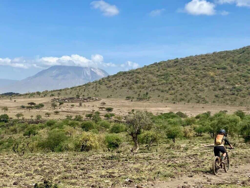 Mountain biker pedaling along rugged singletrack with tall volcano in the distance on the K2N Stage Race Course in Tanzania